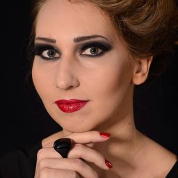 Monika - Portret kobiety z intensywnym makijażem smoky eye, czerwonymi ustami i czarnym pierścieniem na palcu, na ciemnym tle.