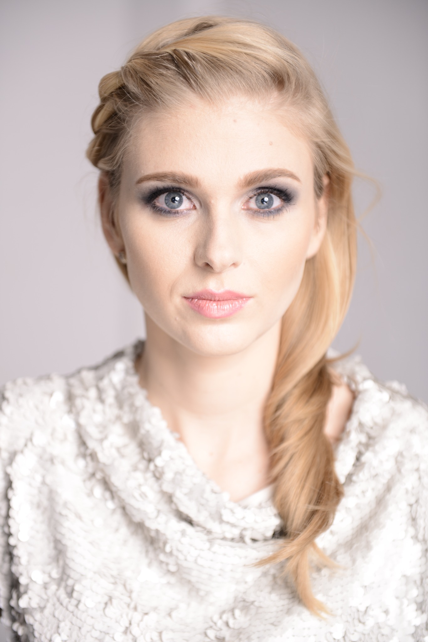 Portret blondwłosej kobiety z warkoczem i subtelnym, wieczorowym podkreśleniem oczu w odcieniach szarości, ubrana w błyszczącą, srebrną bluzkę.