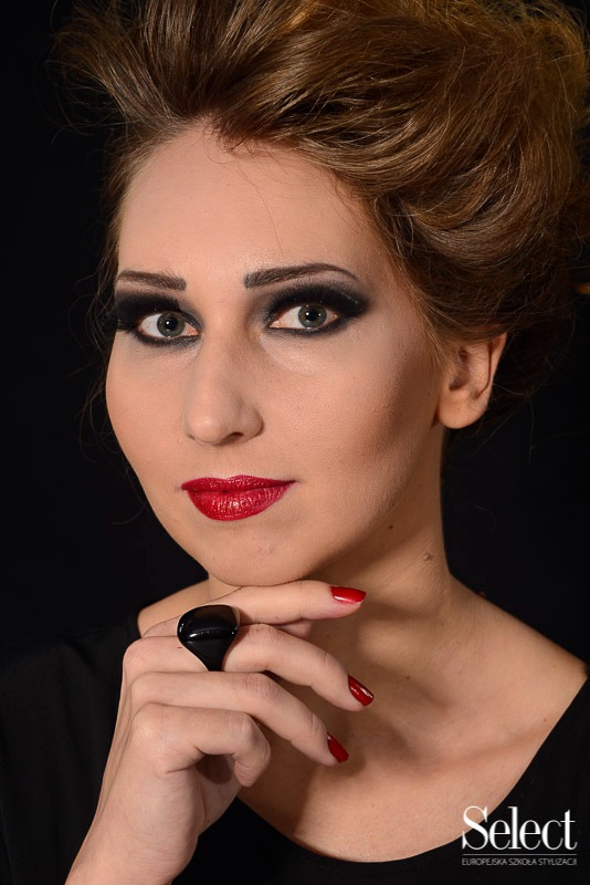 Portret kobiety z intensywnym makijażem smoky eye, czerwonymi ustami i czarnym pierścieniem na palcu, na ciemnym tle.