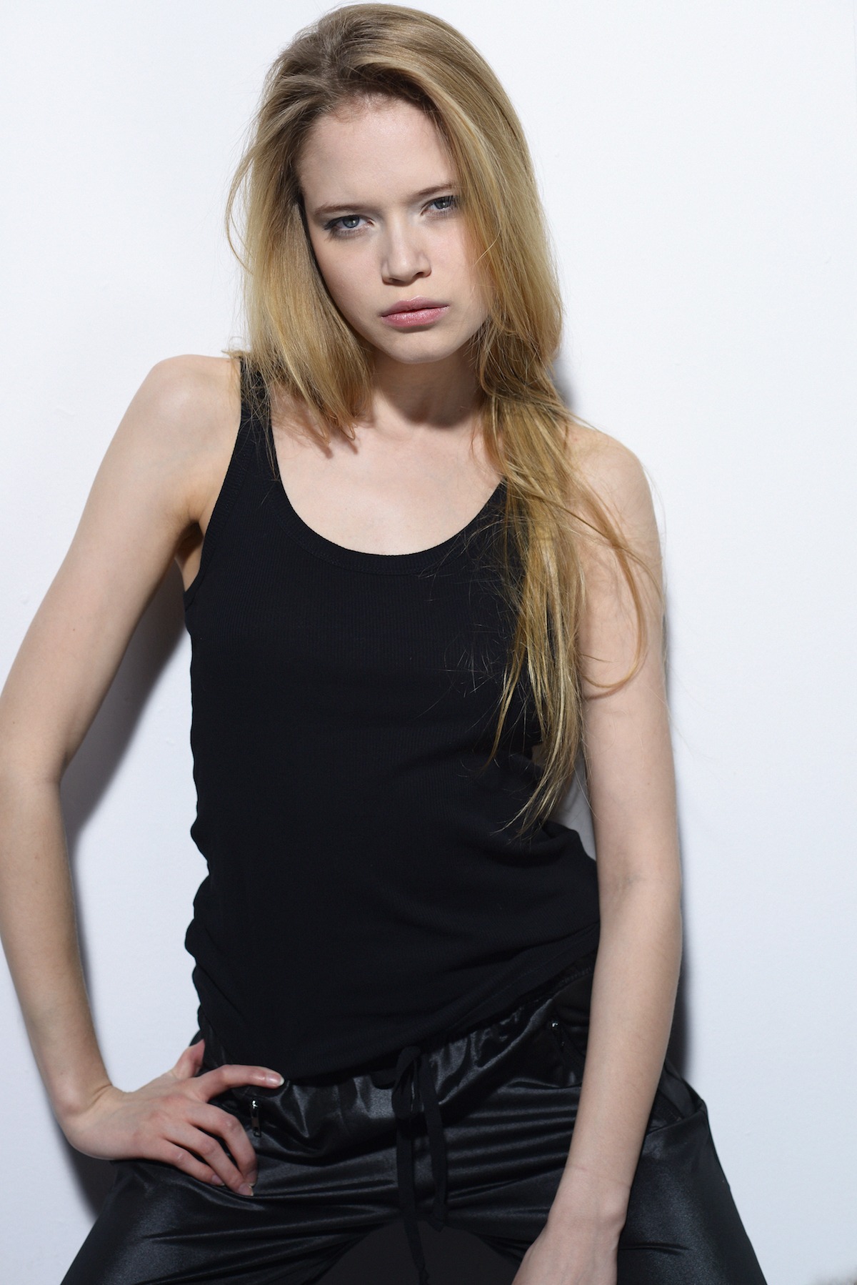 Studyjna fotografia portretowa młodej kobiety z delikatnym, naturalnym upiększeniem, podkreślającym niebieskie oczy i pełne usta, pozującej na tle białej ściany w czarnym topie i satynowych spodniach.