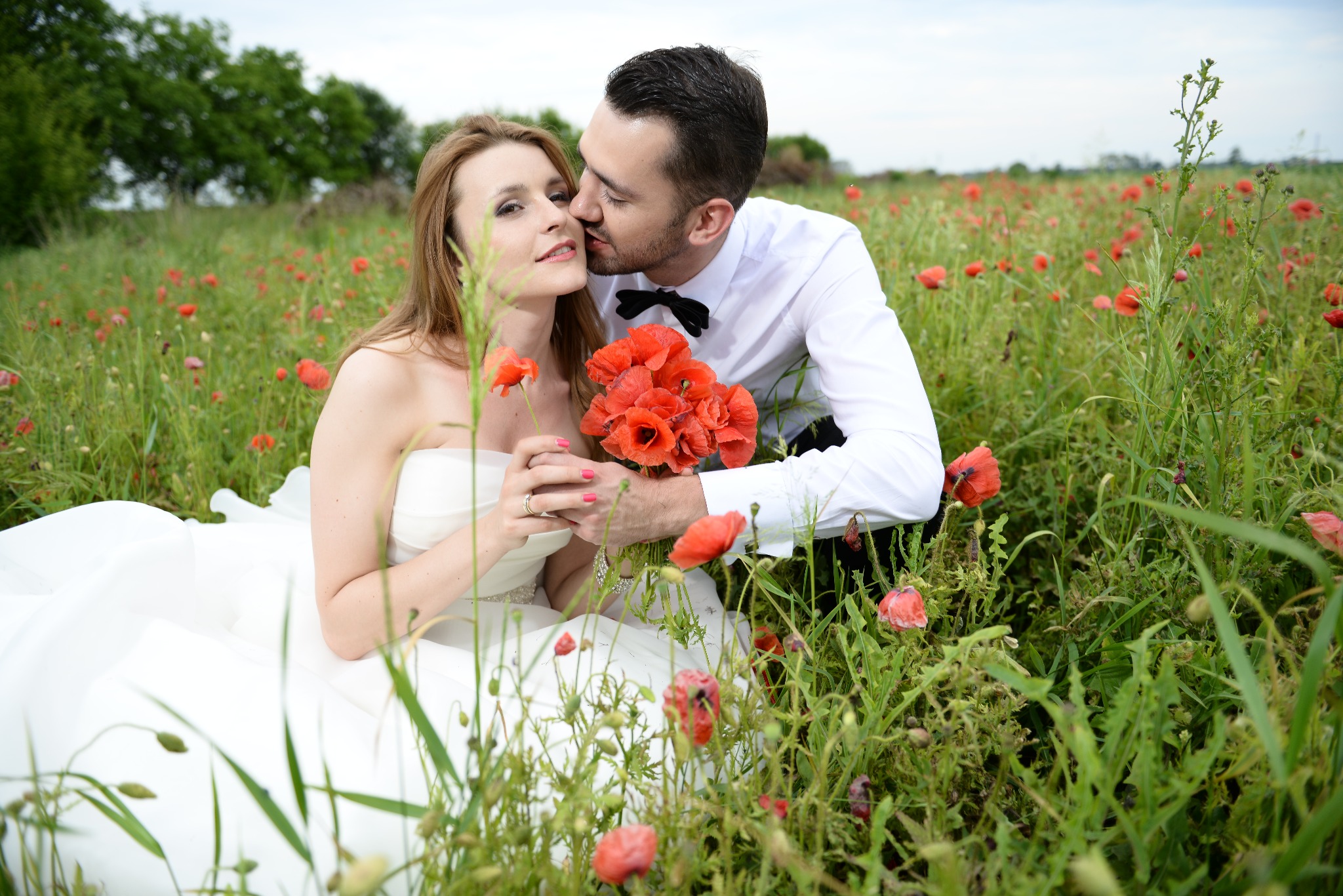 Para młoda w plenerze: panna młoda w białej sukni z bukietem czerwonych maków, pan młody w białej koszuli i muszce całuje ją w policzek, polny krajobraz w tle.