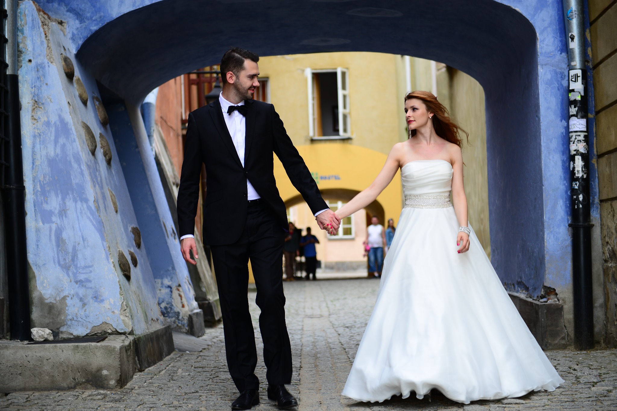 Para młoda trzymająca się za ręce w przejściu z niebieskimi ścianami, panna młoda w białej sukni bez ramiączek z ozdobnym paskiem, pan młody w czarnym smokingu z muszką, w tle budynek z otwartym...