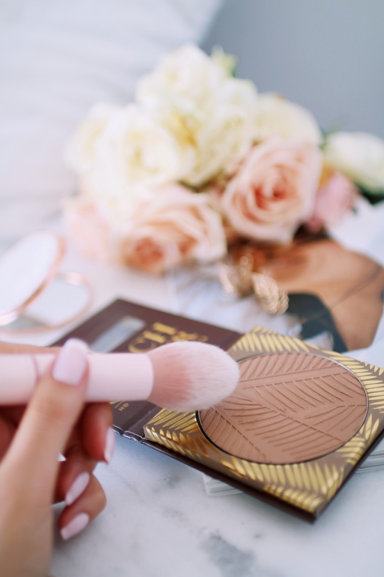 Pudrowy pędzel w dłoni nad bronzerem z wytłaczanym wzorem na tle bukietu róż i lusterka.