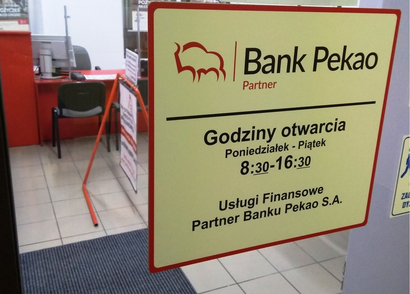 Tablica informacyjna Banku Pekao Partner z godzinami otwarcia (poniedziałek-piątek 8:30-16:30) i logo, widoczna zza drzwi wejściowych, z fragmentem wnętrza oddziału w tle.