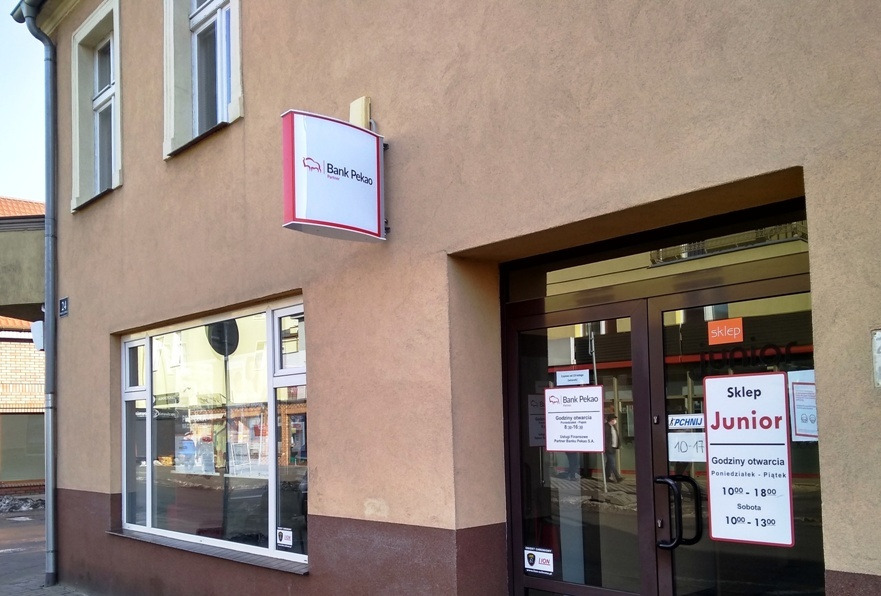Wejście do placówki bankowej Bank Pekao z widocznym szyldem oraz informacją o godzinach otwarcia, obok wejście do sklepu Junior.