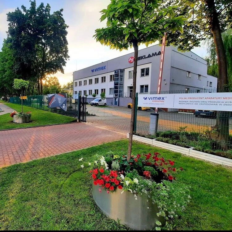 Budynek firmy VIMEX z szarym logo i czerwonym znakiem graficznym na elewacji, widoczny zza metalowego ogrodzenia z tablicą reklamową i ozdobnych donic z czerwonymi i białymi kwiatami.