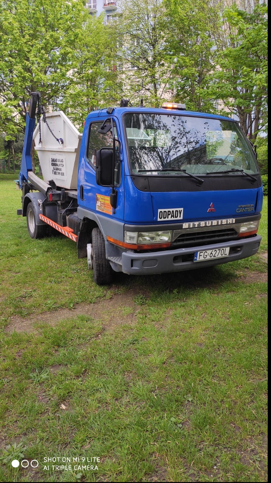 Niebieska ciężarówka Mitsubishi Canter z białym kontenerem na odpady, zaparkowana na trawniku przed budynkiem mieszkalnym. Na kontenerze widoczny napis Zakład Usług Komunalnych i numer telefonu.
