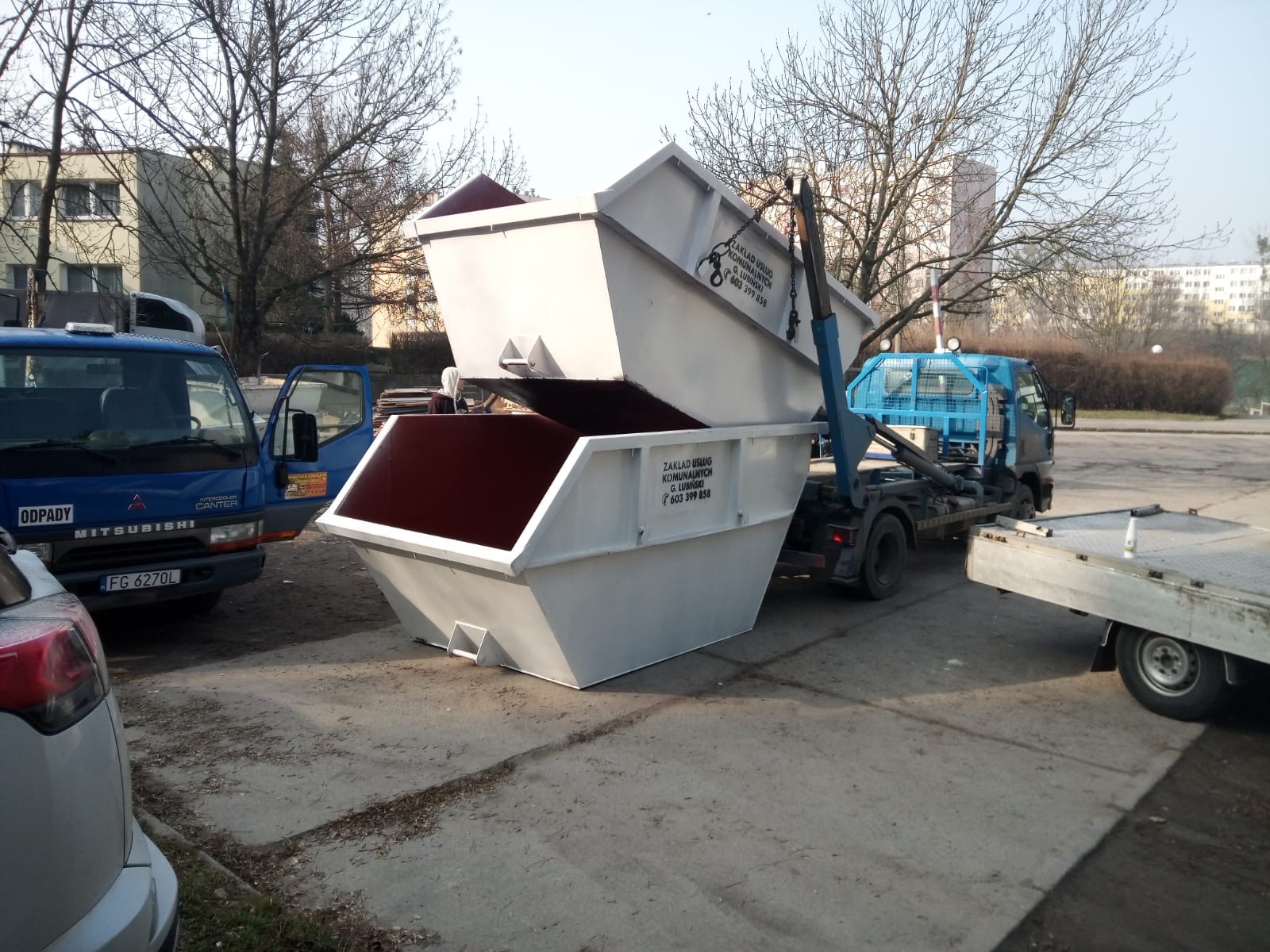 Dwa białe kontenery na gruz, jeden umieszczony na drugim, na tle miejskiego krajobrazu z budynkami i drzewami bez liści, obok zaparkowane samochody i ciężarówka z mechanizmem podnoszącym.