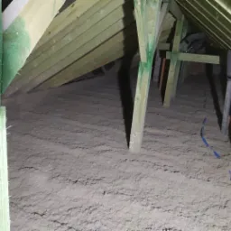 Poddasze z drewnianą konstrukcją pokryte warstwą szarej izolacji, widoczne fragmenty instalacji i pomalowane na zielono elementy konstrukcyjne.