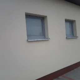 Ocieplona ściana budynku z dwoma szarymi oknami o różnych rozmiarach, widok z zewnątrz, jasny tynk strukturalny.