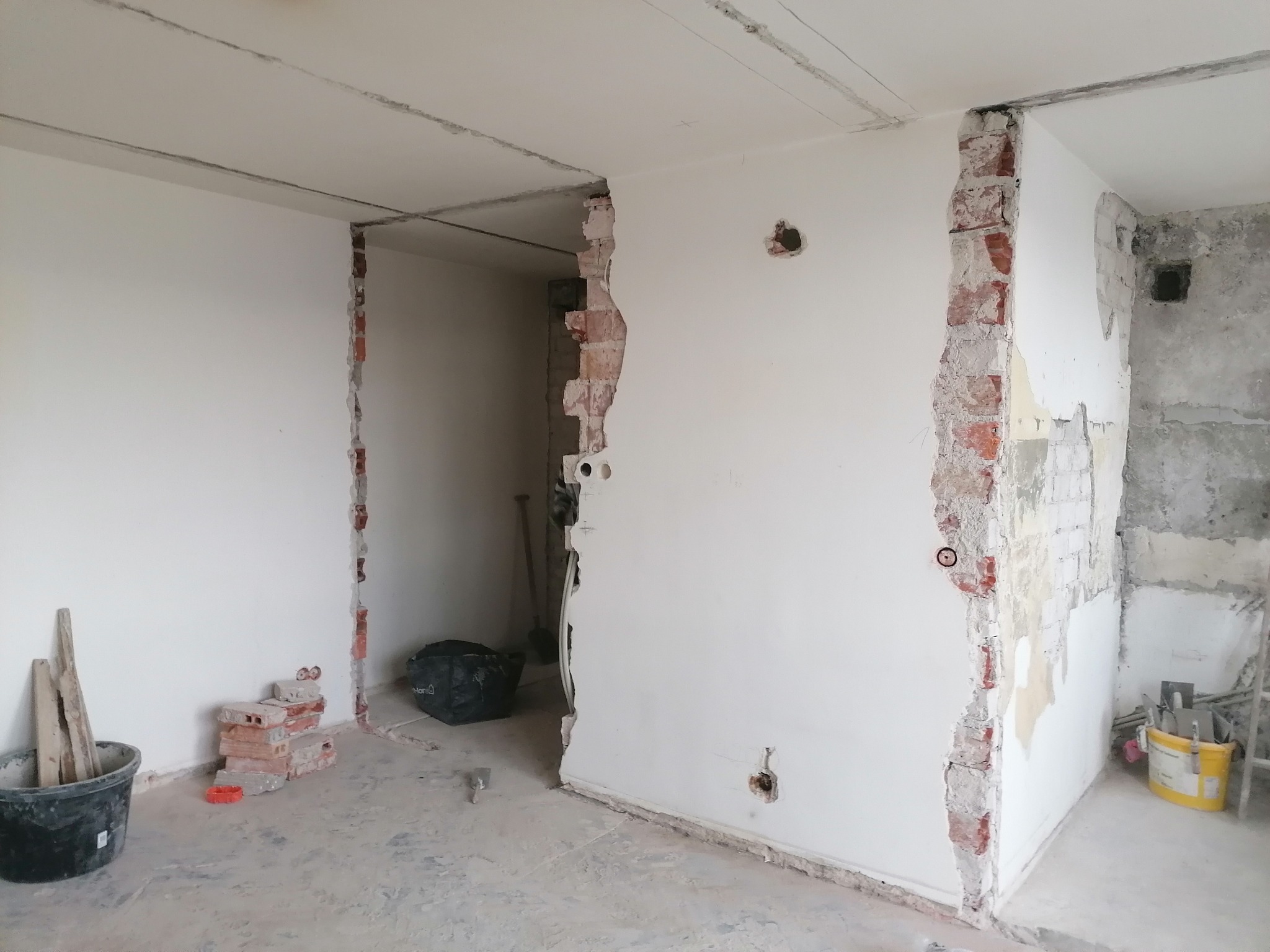 Wnętrze w trakcie remontu: Ściany z częściowo usuniętą warstwą tynku i odsłoniętą cegłą, przygotowane do dalszych prac wykończeniowych. Widoczne narzędzia i materiały budowlane, takie jak wiadro...