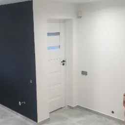 Nowoczesne wnętrze po remoncie: czarna ściana z wystającymi kablami, białe ściany, szare płytki podłogowe z listwami przypodłogowymi, białe drzwi z trzema poziomymi przeszkleniami i czarną klamką...