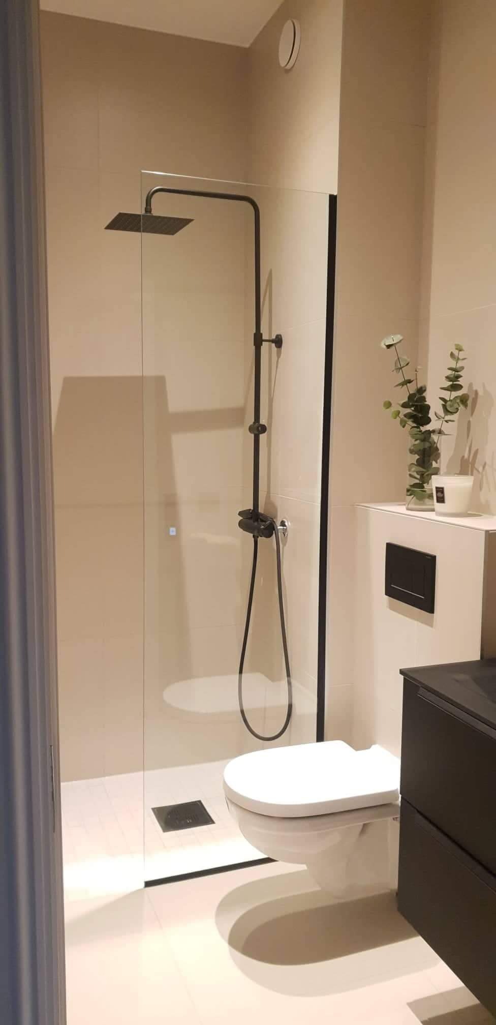 Minimalistyczna łazienka z kabiną prysznicową typu walk-in z czarną armaturą, beżowymi płytkami i wiszącą toaletą, uzupełniona dekoracyjną rośliną w wazonie.