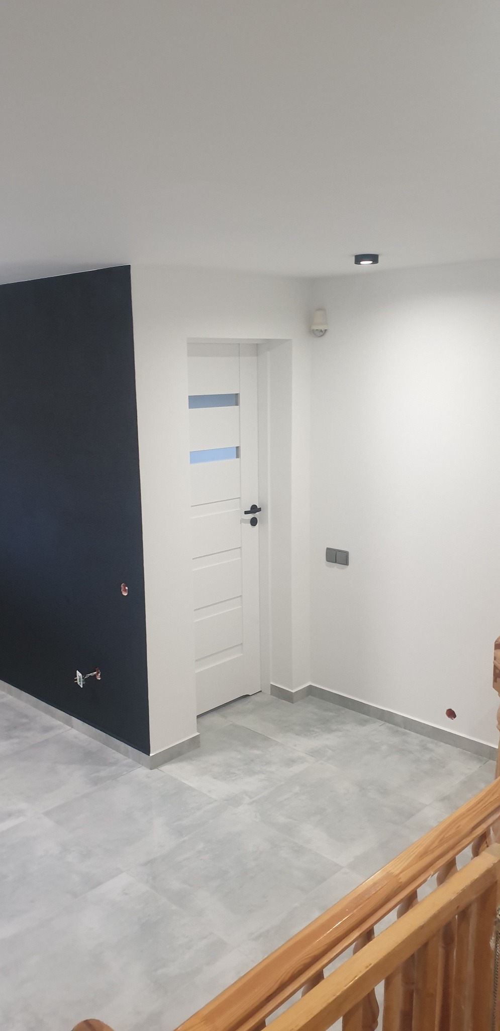 Nowoczesne wnętrze po remoncie: czarna ściana z wystającymi kablami, białe ściany, szare płytki podłogowe z listwami przypodłogowymi, białe drzwi z trzema poziomymi przeszkleniami i czarną klamką...