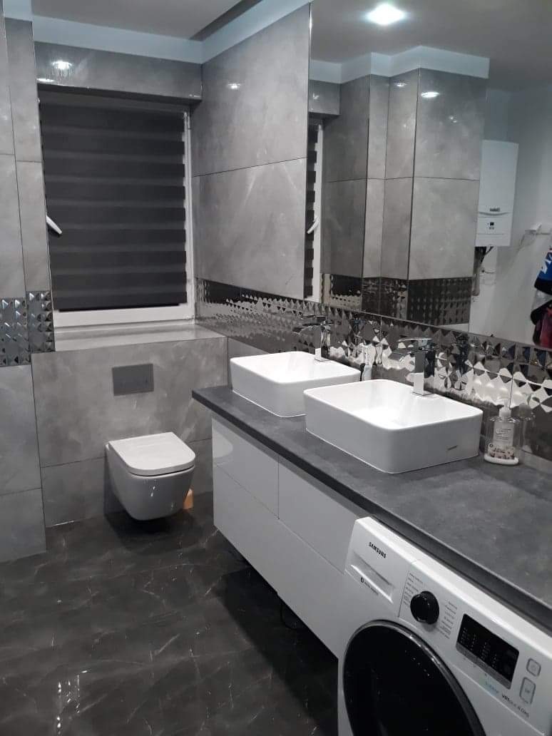 Nowoczesna łazienka wykończona szarymi płytkami imitującymi marmur, z dwiema prostokątnymi umywalkami nablatowymi, toaletą podwieszaną i pralką pod blatem.