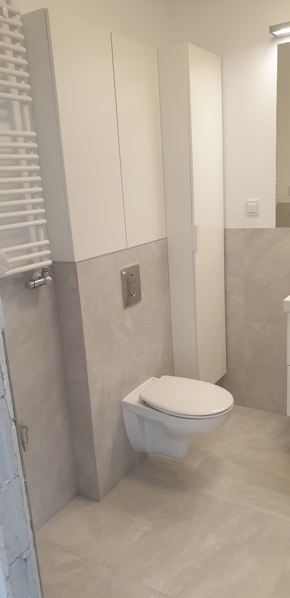 Nowoczesna łazienka z podwieszaną toaletą i grzejnikiem drabinkowym, wykończona płytkami w odcieniach beżu i bieli, z minimalistyczną szafką wiszącą.