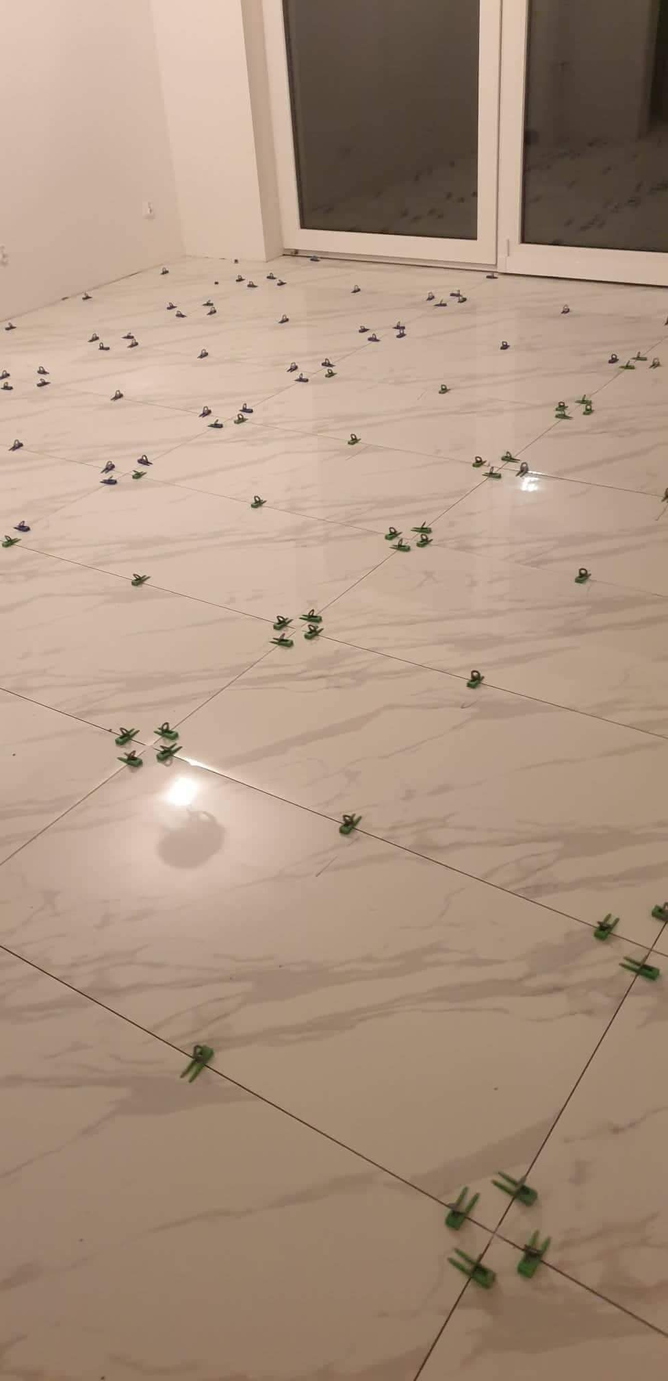 Białe płytki podłogowe w trakcie układania, z widocznymi krzyżykami dystansowymi i systemem poziomowania, w jasnym pomieszczeniu z oknem balkonowym.