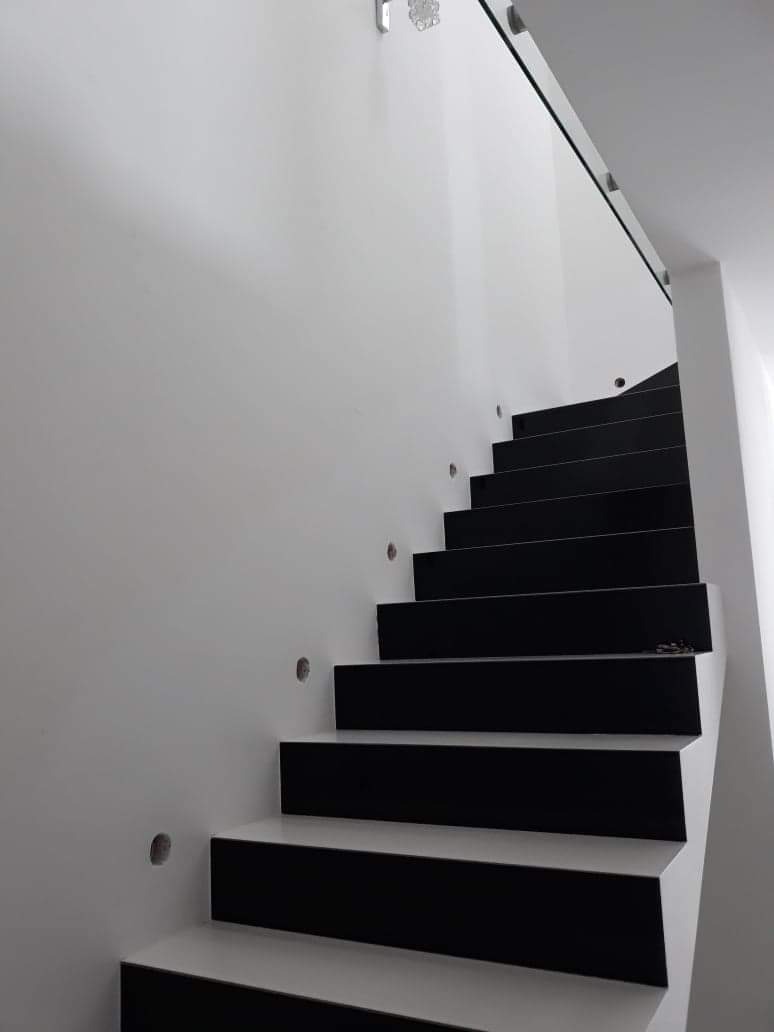 Nowoczesne schody z czarnymi stopniami i białymi podstopnicami, oświetlone punktowymi lampkami w ścianie, zabezpieczone szklaną balustradą.