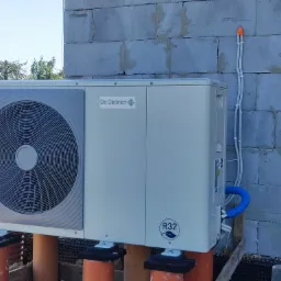 Zewnętrzna jednostka pompy ciepła De Dietrich R32, zainstalowana na pomarańczowych rurach, z widocznymi przewodami w niebieskiej osłonie, przymocowanymi do ściany z szarego betonu komórkowego.