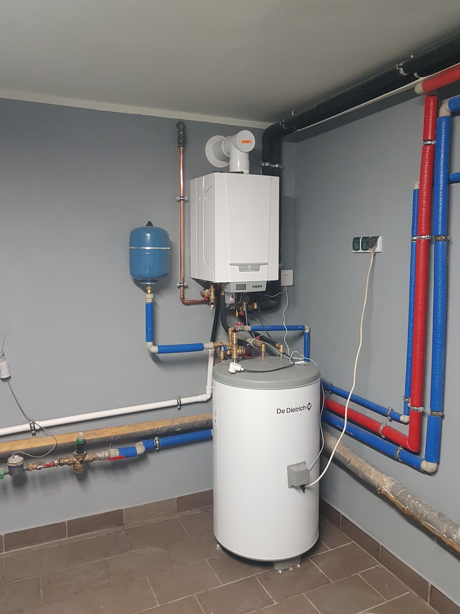 Zestaw grzewczy De Dietrich z piecem gazowym, zasobnikiem ciepłej wody i instalacją hydrauliczną z rurami w kolorach niebieskim i czerwonym, zamontowany w kotłowni z szarą ścianą i płytkami...