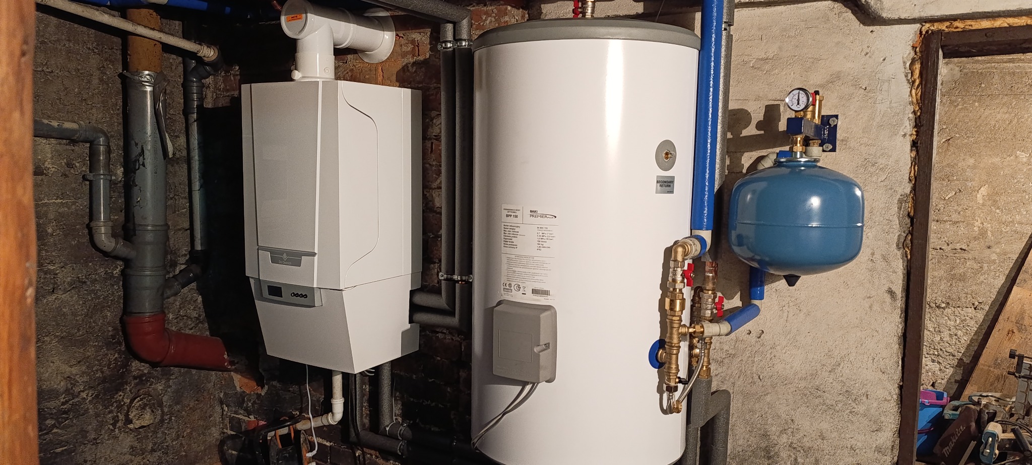 Zestaw instalacji grzewczej: wiszący piec gazowy Baxi, zasobnik ciepłej wody z osprzętem i naczynie przeponowe w piwnicy z widocznymi rurami i starymi ścianami.