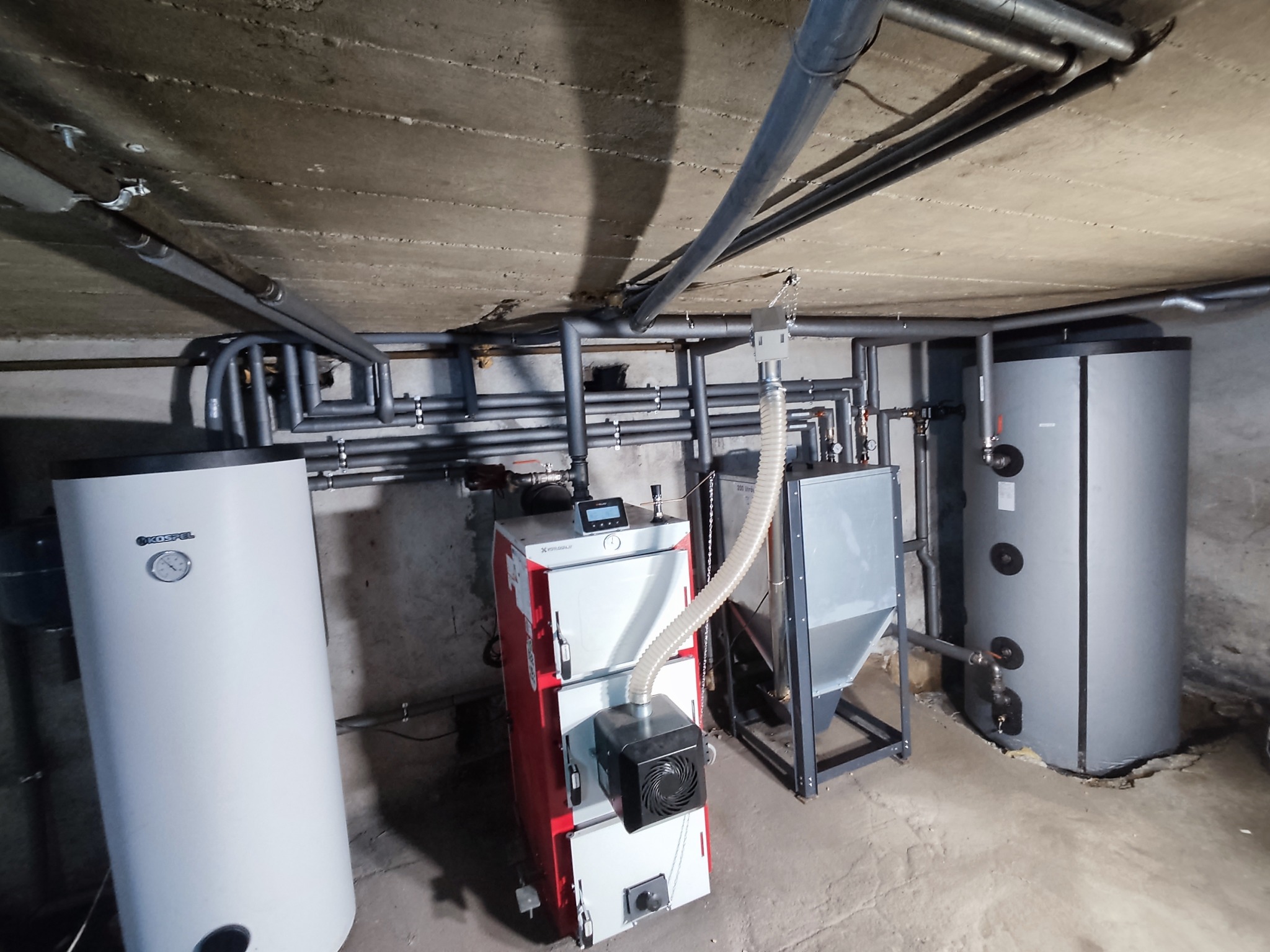 Nowoczesny, automatyczny piec na paliwo stałe (pelet) z podajnikiem, zbiornikiem i dwoma zbiornikami akumulacyjnymi w kotłowni z szarą instalacją grzewczą pod betonowym sufitem.