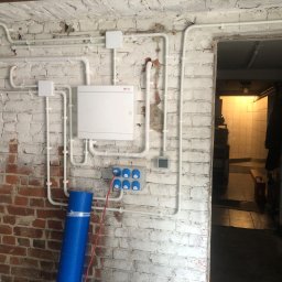 Artur Sałata AS Bud - Nowoczesna instalacja elektryczna z rozdzielnicą i niebieskimi gniazdami siłowymi na częściowo bielonej, surowej ceglanej ścianie w piwnicy lub pomieszczeniu technicznym.