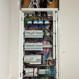 Artur Sałata AS Bud - Uporządkowana instalacja elektryczna w otwartej rozdzielnicy z rzędami wyłączników nadprądowych i różnicówek. Widoczne kolorowe przewody i elastyczne peszle w górnej części wnęki ściennej.
