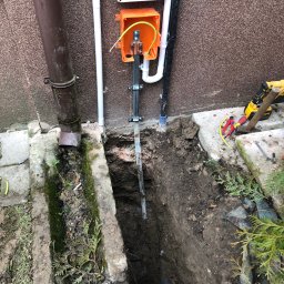 Artur Sałata AS Bud - Zewnętrzna instalacja elektryczna z pomarańczową skrzynką, uziemiającym prętem w świeżo wykopanym rowie obok ściany budynku i brązowej rynny. Widoczne narzędzia.