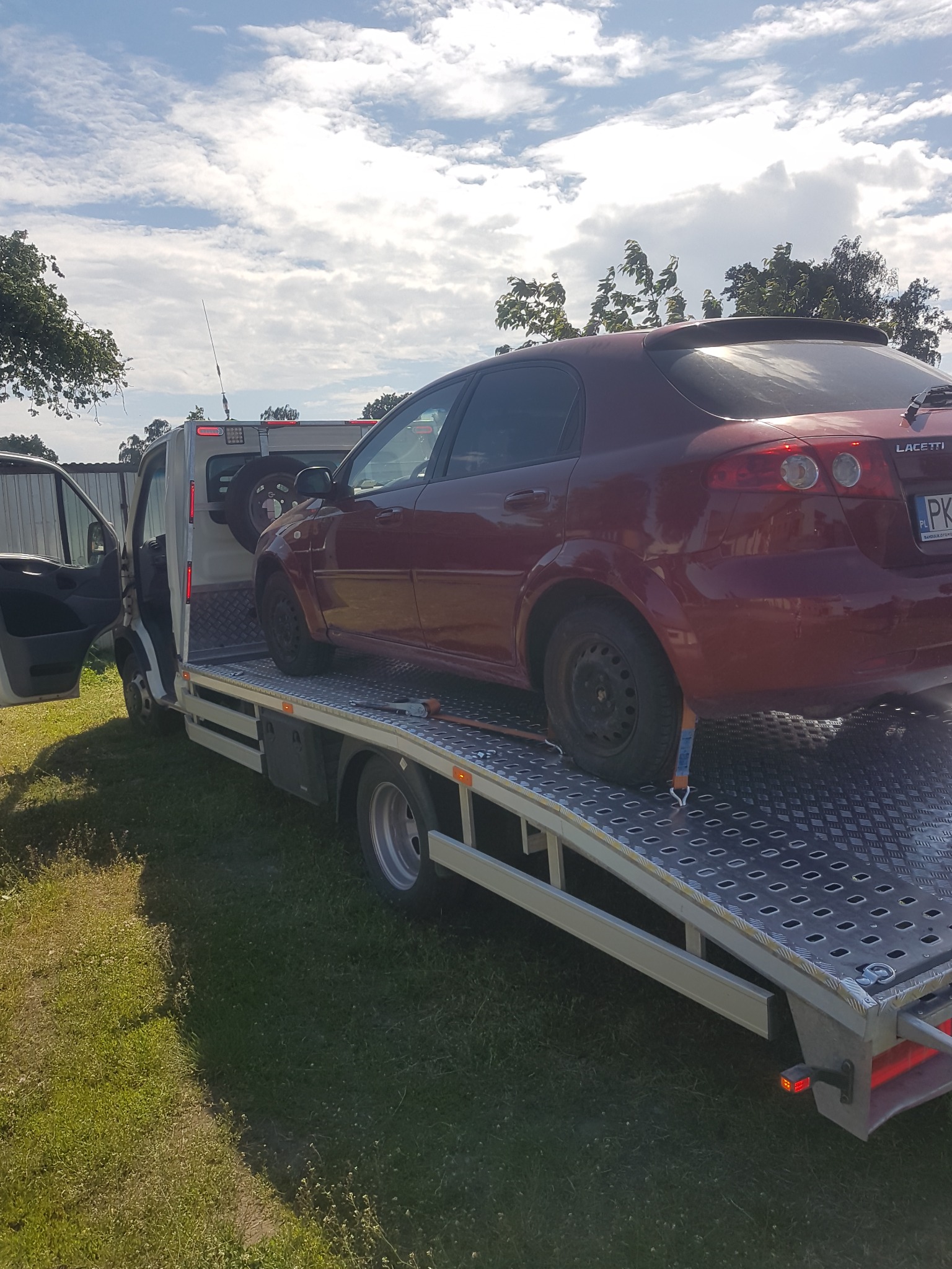 Czerwony samochód osobowy marki Lacetti zabezpieczony pasami na platformie lawety, widok z tyłu, na trawiastym terenie.