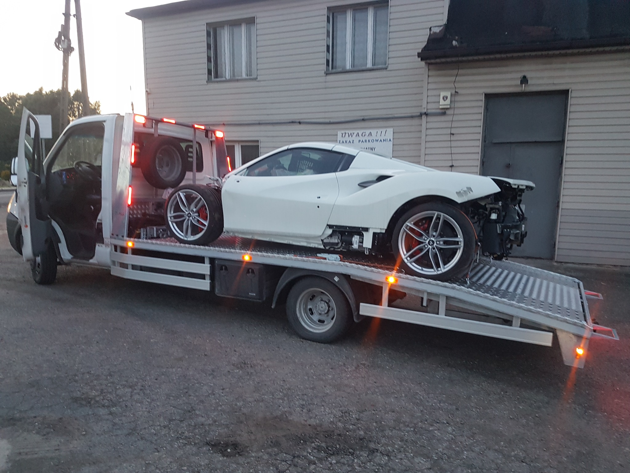 Laweta z otwartymi drzwiami przewozi uszkodzony, biały samochód sportowy bez przedniego zderzaka i z dodatkowymi kołami z tyłu, zaparkowana na tle budynku z szarą elewacją i znakiem 'Uwaga! Zakaz...