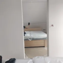 Widok przez ościeżnicę na łóżko z drewnianą ramą i materacem w folii, w tle poddasze i osoba robiąca zdjęcie telefonem.