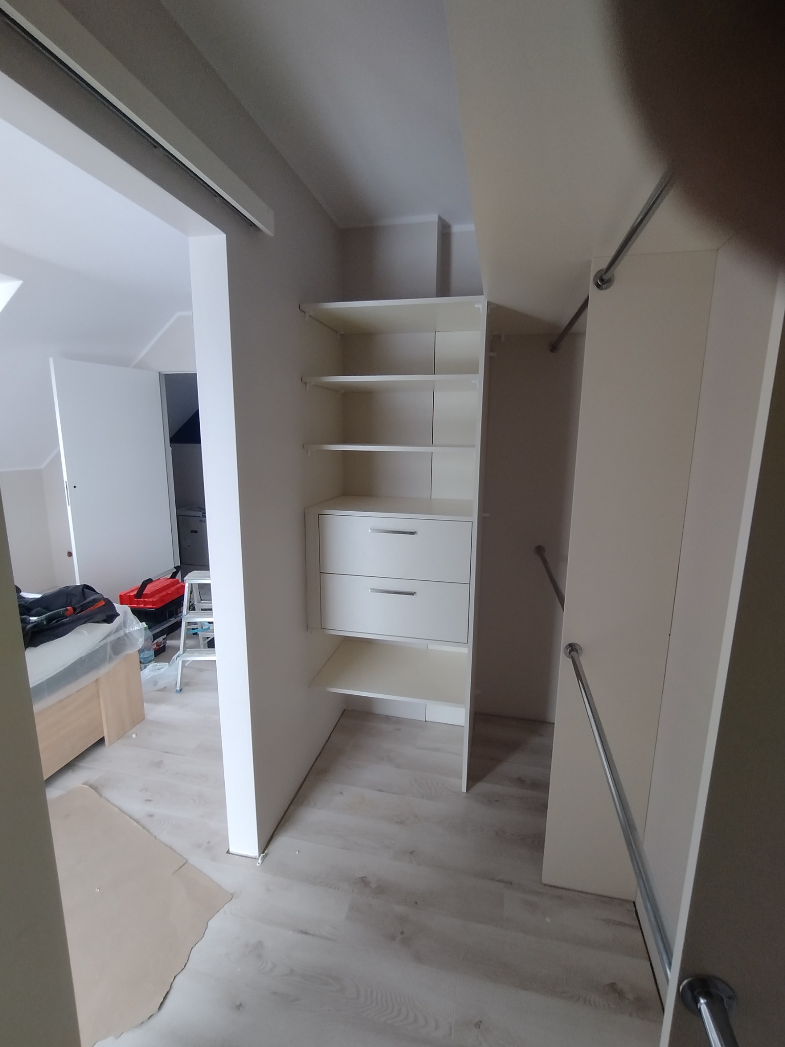 Montaż szafy wnękowej: widok na wnętrze szafy z półkami i szufladami, w tle widoczne narzędzia i elementy wyposażenia pokoju, podłoga częściowo zabezpieczona kartonem.