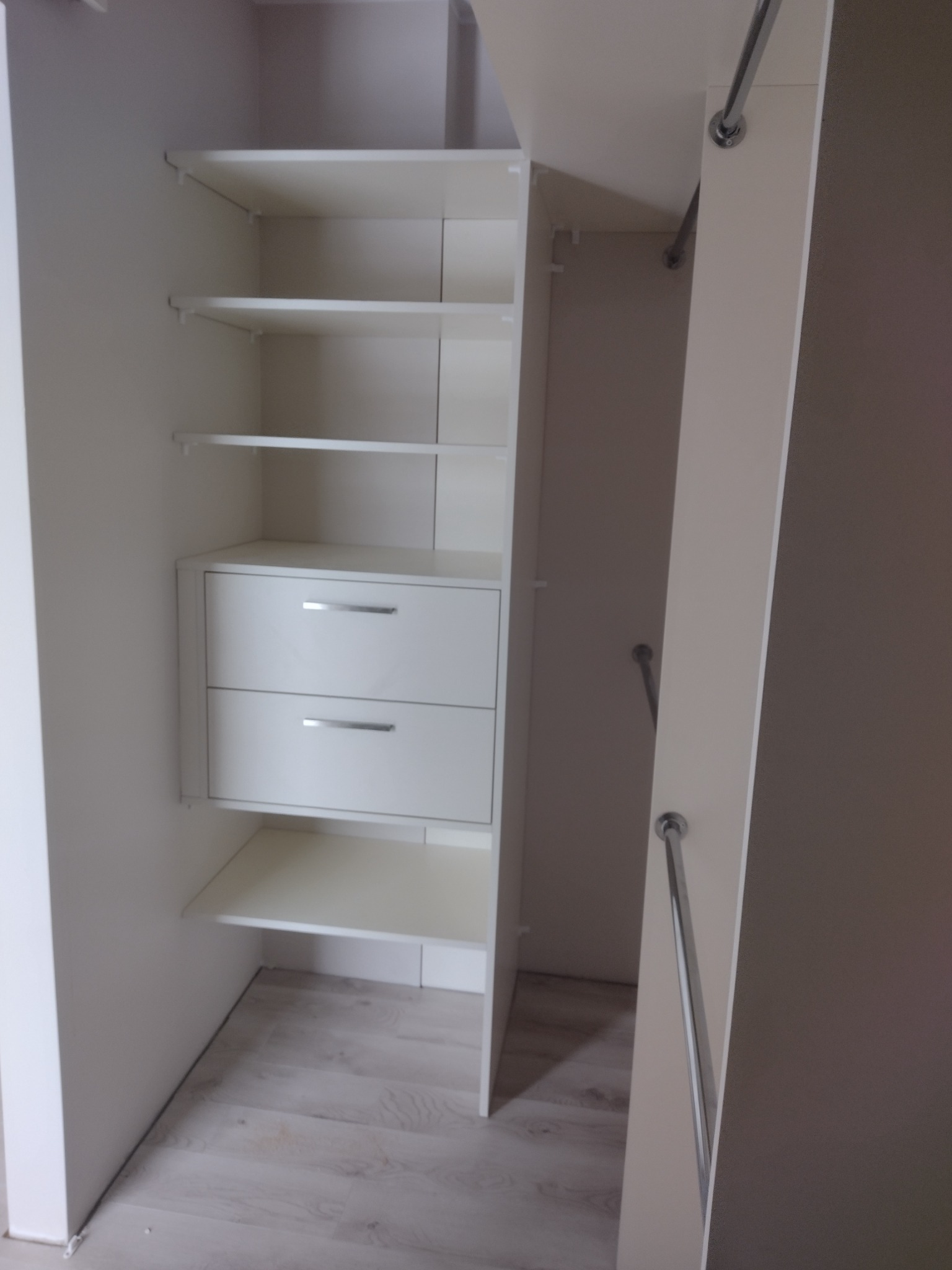 Wnętrze białej szafy wnękowej z półkami i dwiema szufladami z metalowymi uchwytami, widoczne jasne panele podłogowe.