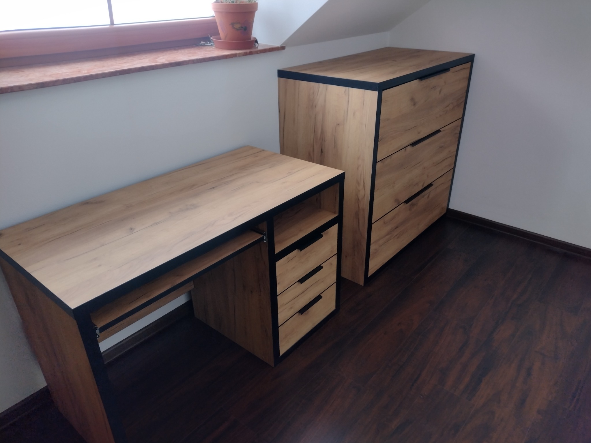 Komplet mebli do pokoju na poddaszu: biurko z szufladami i wysoka komoda z drewnopodobnym fornirem i czarnymi obramowaniami, ustawione na ciemnej podłodze.