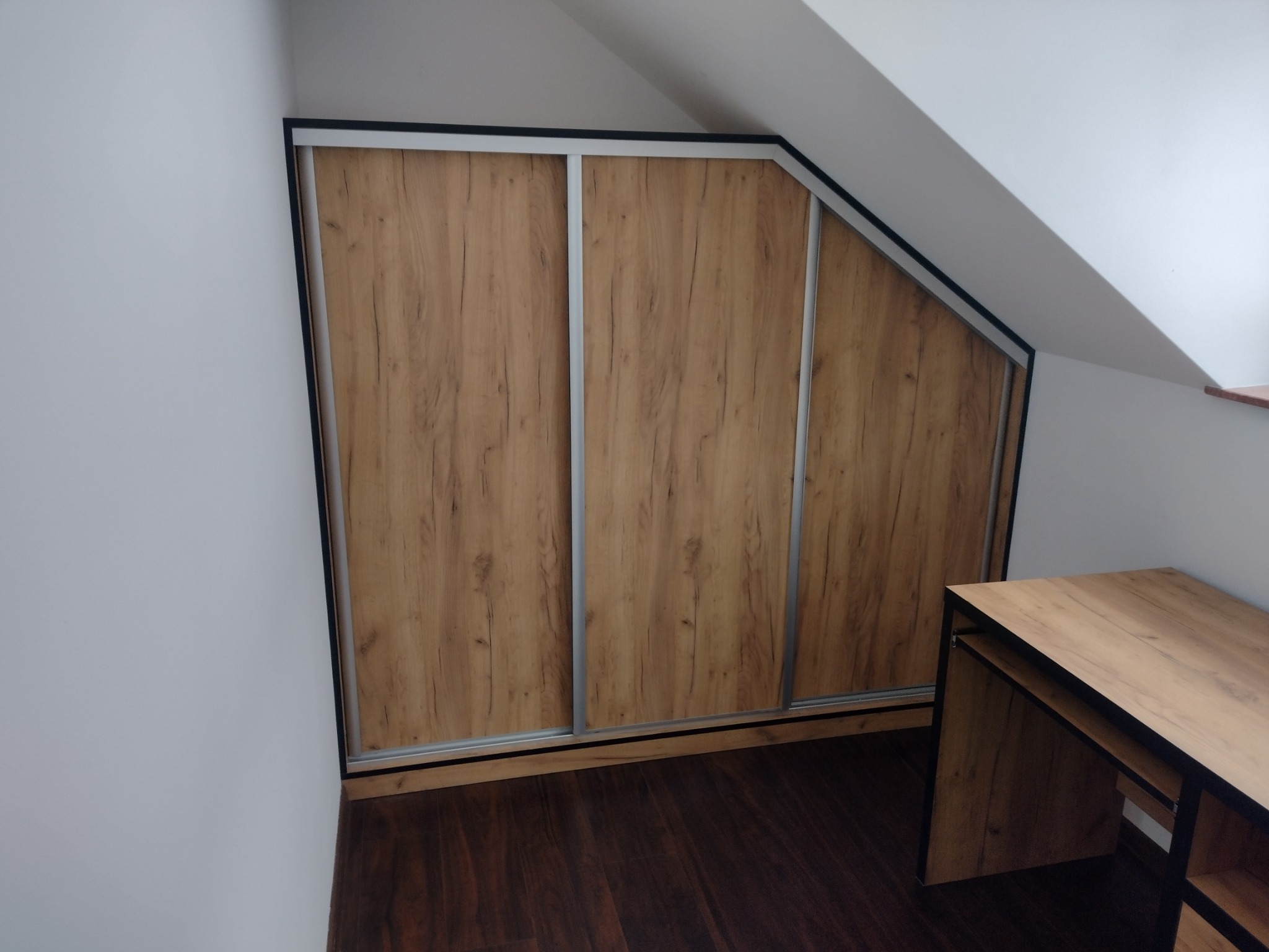 Trzydrzwiowa szafa wnękowa z przesuwnymi drzwiami o fakturze drewna, dopasowana do skosu poddasza, z blatem biurka w podobnej tonacji.