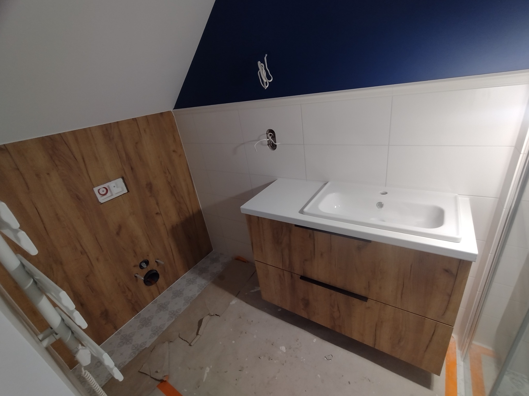 Łazienka w trakcie remontu: szafka pod umywalkę z drewnopodobnym frontem, biały blat i umywalka, białe płytki na ścianie, niebieska ściana na poddaszu, widoczne przewody elektryczne, grzejnik...