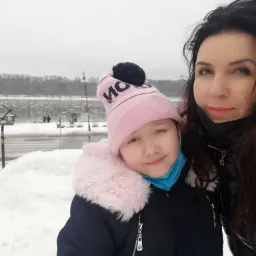 Portret matki i córki w zimowej scenerii; dziewczynka w różowej czapce i niebieskiej maseczce, kobieta w czarnej kurtce i maseczce, w tle ośnieżony krajobraz z rzeką i przechodniami.