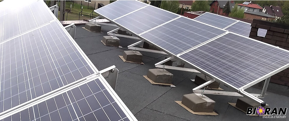 Panele słoneczne na płaskim dachu, zamontowane na betonowych podstawach, widok z góry na tle zabudowań.