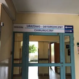 Tablica informacyjna z napisem 'Oddział Urazowo-Ortopedyczny Chirurgiczny' nad wejściem do oddziału szpitalnego, strzałki wskazujące kierunek, widoczne fragmenty wnętrza oddziału przez przeszklone...