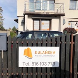 ART-REKLAMA - Tablica reklamowa z logo firmy Kulańska Nieruchomości umieszczona na drewnianym ogrodzeniu przed budynkiem biurowym, widoczna skrzynka pocztowa po lewej stronie.