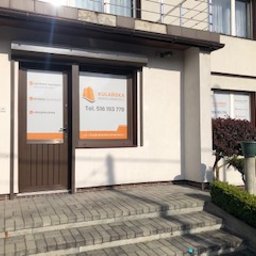 ART-REKLAMA - Fasada budynku z witryną i wejściem, na których umieszczono pomarańczowe grafiki informacyjne z danymi kontaktowymi firmy.