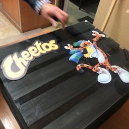 ART-REKLAMA - Naklejka z logo Cheetos i postacią geparda umieszczona na czarnym, drewnianym pudełku. Widoczna ręka osoby umieszczającej naklejkę.