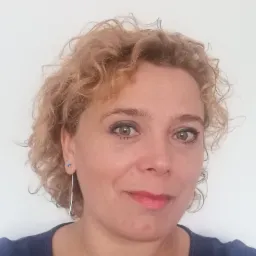 Portret kobiety z kręconymi blond włosami, ubrana w granatową bluzkę, na białym tle.