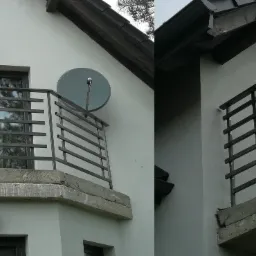 Szara, metalowa balustrada balkonowa z poziomymi prętami na betonowej płycie balkonu, widoczna antena satelitarna i lampa zewnętrzna na ścianie budynku.