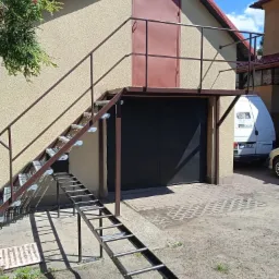 Zewnętrzne schody metalowe z platformą prowadzące do drzwi na piętrze garażu, dodatkowa drabinka metalowa na dole, widok z dołu.