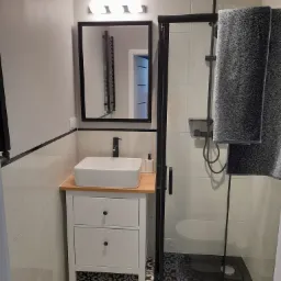 Nowoczesna łazienka z kabiną prysznicową typu walk-in z czarnymi ramami, umywalką nablatową na szafce z szufladami i lustrem w czarnej ramie, wykończona płytkami w odcieniach bieli i wzorzystą...