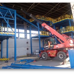 Larget Sp. z o.o. - Montaż niebieskiej stalowej konstrukcji hali z żółtymi barierkami, widoczny czerwony wózek widłowy z operatorem w pomarańczowej kamizelce.