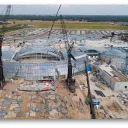 Budowa hali z przeszklonym dachem kopułowym, widok z drona, kilka dźwigów w trakcie pracy, porozrzucane elementy konstrukcyjne na placu budowy.