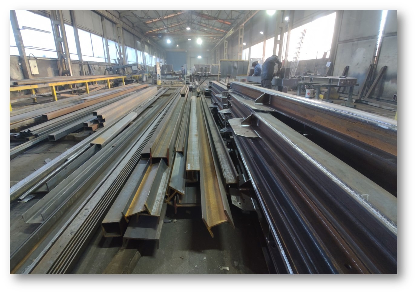 Stalowe profile o różnych przekrojach ułożone w hali produkcyjnej, widoczne ślady spawania na niektórych elementach, w tle pracownicy przy stanowiskach.